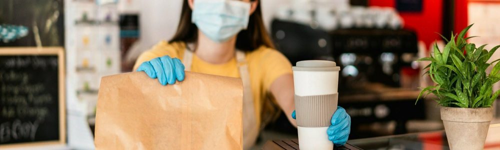 Young woman wearing face mask while serving takeaway breakfast and coffee inside cafeteria restaurant - Worker preparing healthy food inside cafè bar during coronavirus period - Focus on right hand